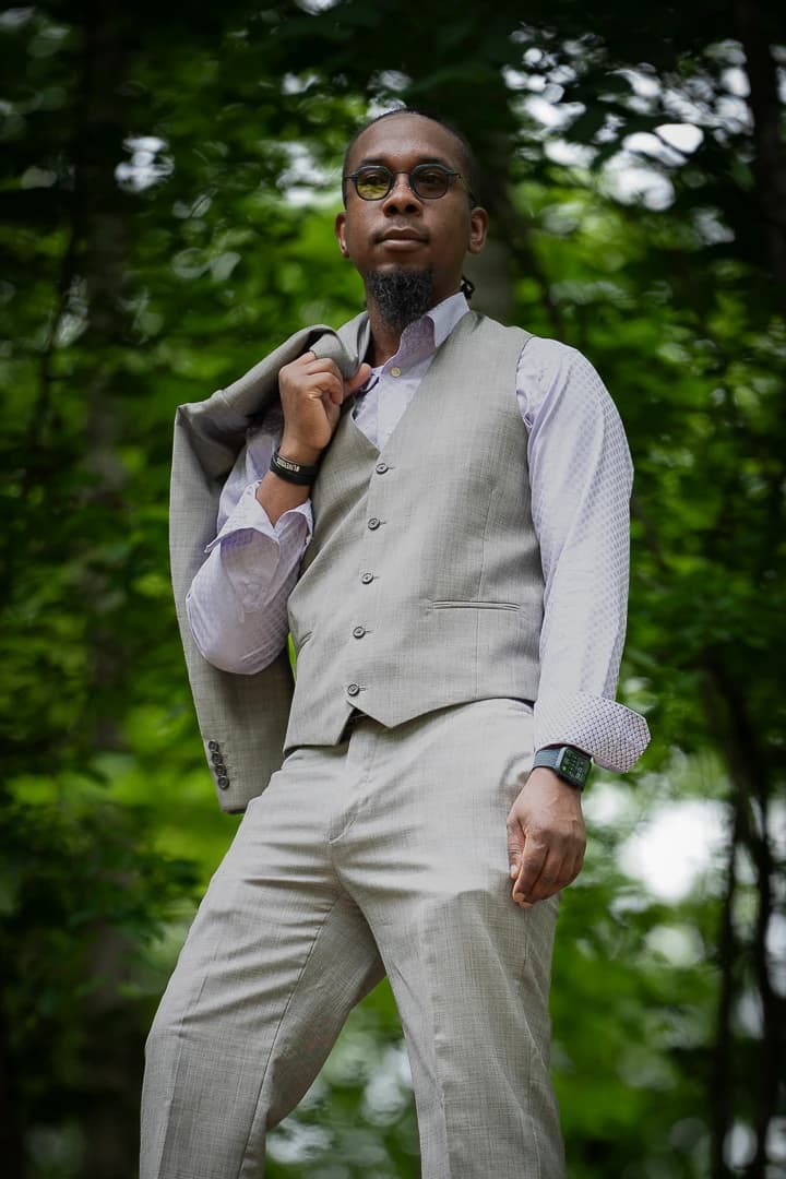 Man in round tinted glasses and a grey three-piece suit facing the camera in a green forest