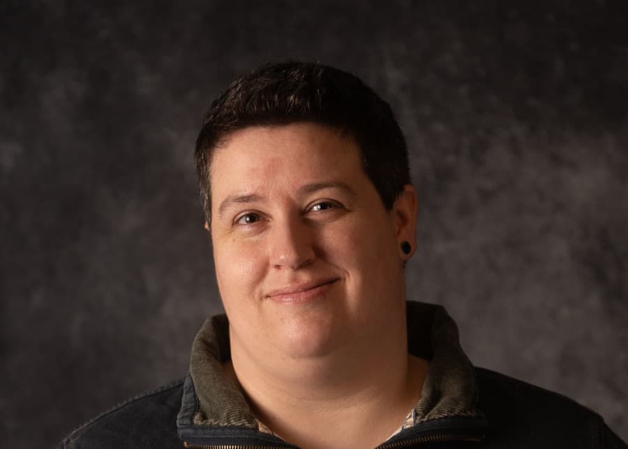 Smiling person with short dark hair in a striped pullover against a grey studio backdrop
